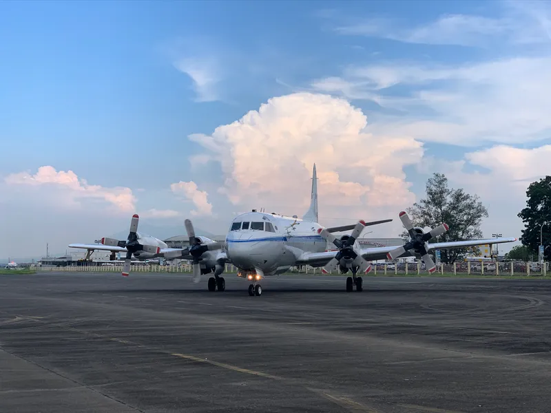 Image of a NASA P-3 Orion aircraft on the tarmac with a large storm in the background.